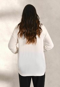White long-sleeve shirt with a smooth texture, rounded hem, and lightweight fabric, paired with black pants. Back view shows flowing hairstyle.