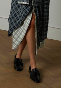 Zwarte loafers met een glanzende afwerking, voorzien van een groot stoffen strikdetail aan de voorkant. De zool is slank en de schoenen hebben een lage hak.