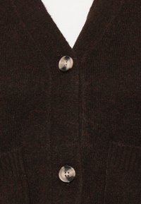 Cardigã tricotado castanho-escuro com dois botões grandes em mármore, preso sobre uma blusa branca com nervuras.