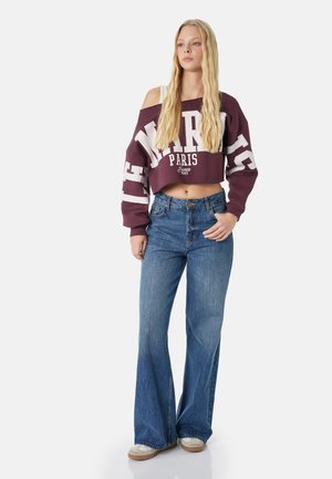 Junge Frau mit langen blonden Haaren, die ein bordeauxfarbenes, kurz geschnittenes Off-Shoulder-Sweatshirt mit dem Schriftzug „Paris“, blaue ausgestellte Jeans und lässige Sneaker trägt.
