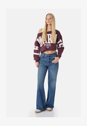 Jonge vrouw met lang blond haar, die een bordeauxrood cropped off-shoulder sweatshirt met de tekst "Paris" draagt, blauwe uitlopende spijkerbroek en casual sneakers.