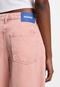 Vaaleanpunaiset farkut, joissa on sininen HUGO-merkkinen kangasmerkki vyötäröllä, yksittäinen takatasku ja hienovaraiset tekstuurielementit.