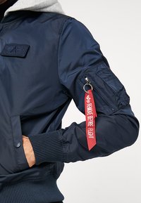 Chaqueta bomber azul marino con capucha gris, que cuenta con un bolsillo en el brazo con cremallera y una etiqueta de tela roja etiquetada "RETIRAR ANTES DEL VUELO". Puños y dobladillo acanalados.