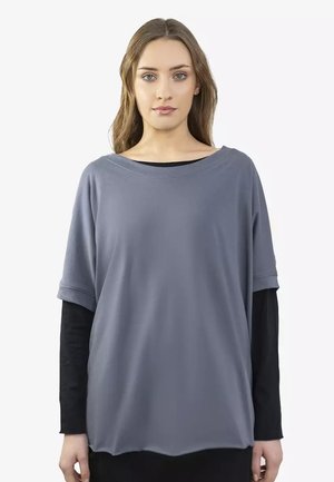 Kobieta o długich brązowych włosach, ubrana w luźny szary top z krótkim rękawem, nałożony na dopasowaną czarną koszulę z długim rękawem, zwrócona twarzą do przodu z neutralnym wyrazem.