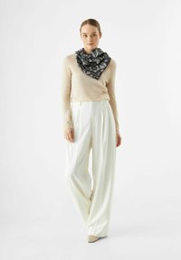 Lichtbeige trui, hooggetailleerde witte broek, zwart dessin sjaal geknoopt in een driehoekvorm, schoenen in neutrale tinten. Eenvoudig, elegant ontwerp.