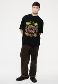 Black oversized t-shirt featuring a colorful graphic design with "Fleetwood Mac" text, paired with brown corduroy pants and black shoes.