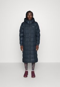 Lang polstret frakk, mørk marinblå, med hette, glidelås og elastiske mansjetter. Sammen med mønstrede leggings og burgunder sneakers.