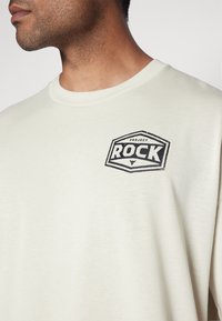 Beige bavlněné tričko s kulatým výstřihem a krátkými rukávy. Na levé straně hrudi se nachází černé logo "PROJECT ROCK".