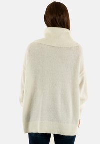 Witte oversized trui met een hoge, geribde kraag, gemaakt van een zachte, gestructureerde stof en een losse pasvorm, gecombineerd met donkere denimjeans.