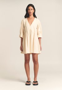 Lichtbeige katoenen jurk met een V-hals, driekwart pofmouwen en een geraffineerde taille; boven de knie lengte met een losse pasvorm.
