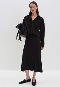 Donna che indossa una blusa nera a maniche lunghe e una gonna midi, tiene in mano una borsa nera strutturata, in piedi davanti a uno sfondo bianco uniforme.