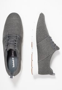 Šedé textilní boty s texturovaným svrškem, kulatou špičkou a šněrováním. Bílá gumová podrážka s hnědými akcenty. Obsahuje poutko na patě.