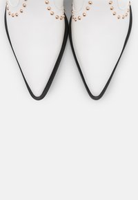 Zapatos blancos de punta afilada con suelas negras, acentuados por tachuelas de oro rosa dispuestas en un patrón circular cerca de las aberturas. Textura suave.