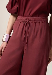 Bordeaux, lichte, wijde broek met een elastische tailleband en een trekkoorddetail. Gecombineerd met een geometrische, veelkleurige armband.