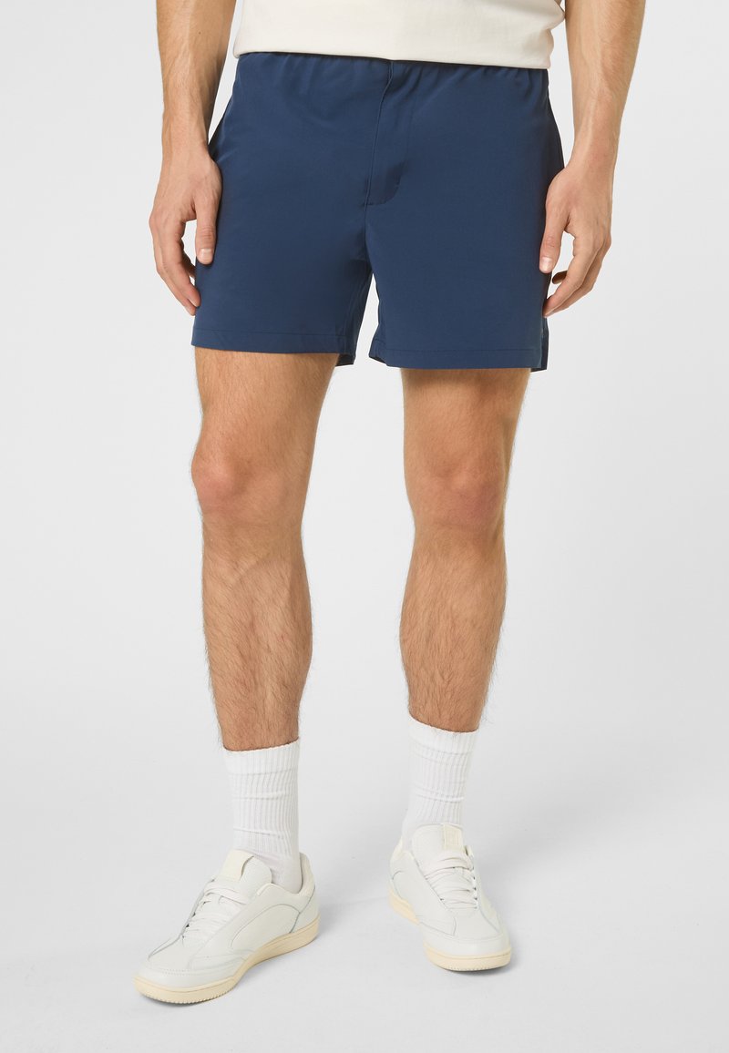 Homme portant un short de sport bleu marine, des chaussettes blanches montantes et des baskets blanches, debout devant un fond clair uni.