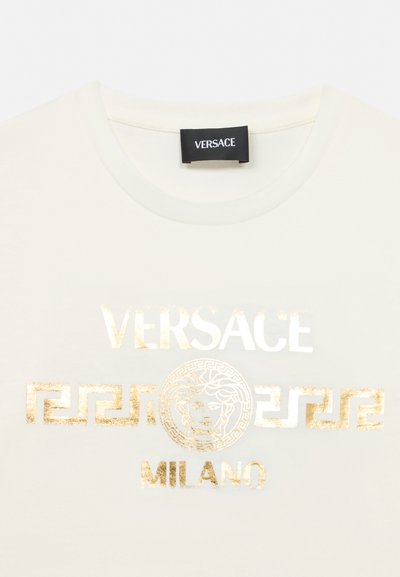 Tricou alb cu decolteu rotund, cu text auriu "VERSACE MILANO" și logo-ul capului Medusa centrat pe piept. Etichetă neagră Versace pe gât.