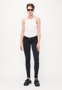 Črne skinny kavbojke s prilegajočim dizajnom, v kombinaciji z belo brezrokavno majico. Vključujejo robustne črne čevlje in pravokotne sončne očala.
