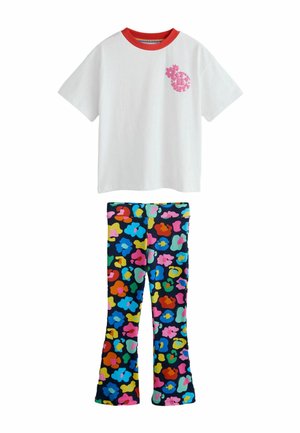 Lachend kind met kort bruin haar draagt een wit oversized T-shirt en kleurrijke patroonbroek, staand op een neutrale achtergrond.