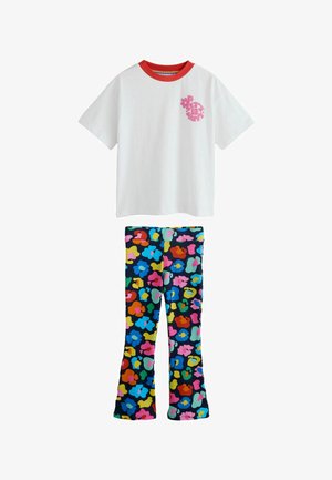 Lachend kind met kort bruin haar draagt een wit oversized T-shirt en kleurrijke patroonbroek, staand op een neutrale achtergrond.