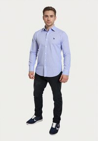 Giovane uomo in piedi, che indossa una camicia a righe blu chiaro con bottoni, pantaloni neri e sneakers blu navy con lacci bianchi.
