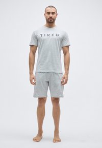 Γκρι βαμβακερό t-shirt με την επιγραφή «TIRED», συνδυασμένο με αντίστοιχα γκρι βαμβακερά σορτς. Μινιμαλιστικός σχεδιασμός, απαλή υφή, φορεμένα ξυπόλητα.