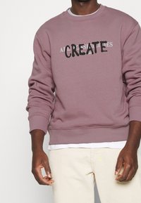 Sweatshirt violet avec le texte "CREATE" en noir, col rond, poignets et ourlet côtelés, associé à un pantalon en velours côtelé beige clair.