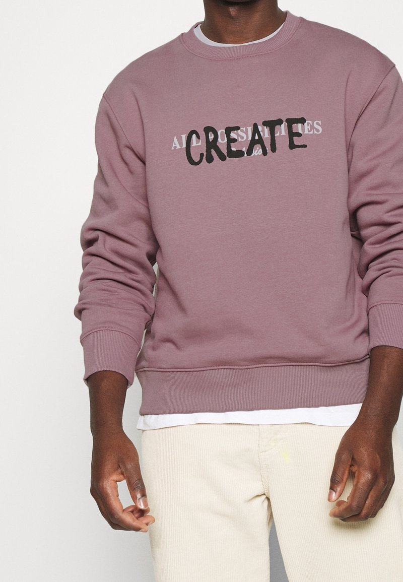 Sweatshirt violet avec le texte "CREATE" en noir, col rond, poignets et ourlet côtelés, associé à un pantalon en velours côtelé beige clair.