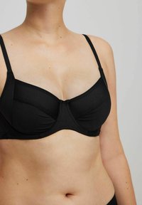 Acercamiento de una persona que lleva un sujetador negro con aros y tirantes finos frente a un fondo gris claro liso.