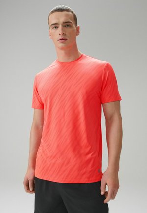 Junger Mann steht, trägt ein kurzärmeliges Sportshirt mit Korallenmuster und schwarze Shorts vor einem schlichten grauen Hintergrund.