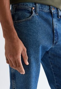 Close-up af en persons venstre hånd hvilende ved siden af blå denimjeans med synlige stikninger og kobbernitter.