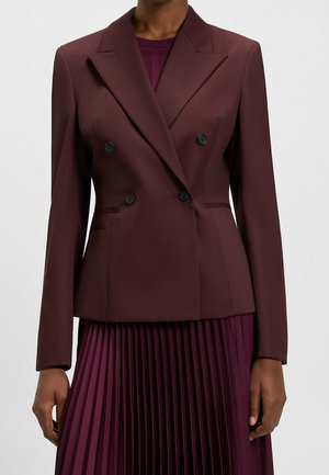 Mujer vistiendo un blazer burdeos entallado de doble botonadura sobre una falda plisada a juego, de pie frente a un fondo liso.