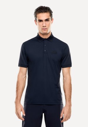 Giovane con i capelli pettinati all'indietro, indossa una polo blu navy aderente e pantaloni abbinati, in piedi su uno sfondo chiaro e semplice.