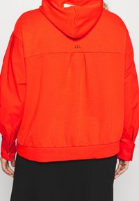 Červená mikina s kapucí a volným střihem, s plisovaným zadním dílem, panelovým detailem a knoflíkovými ozdobami na manžetách. Na zádech je potisk loga.