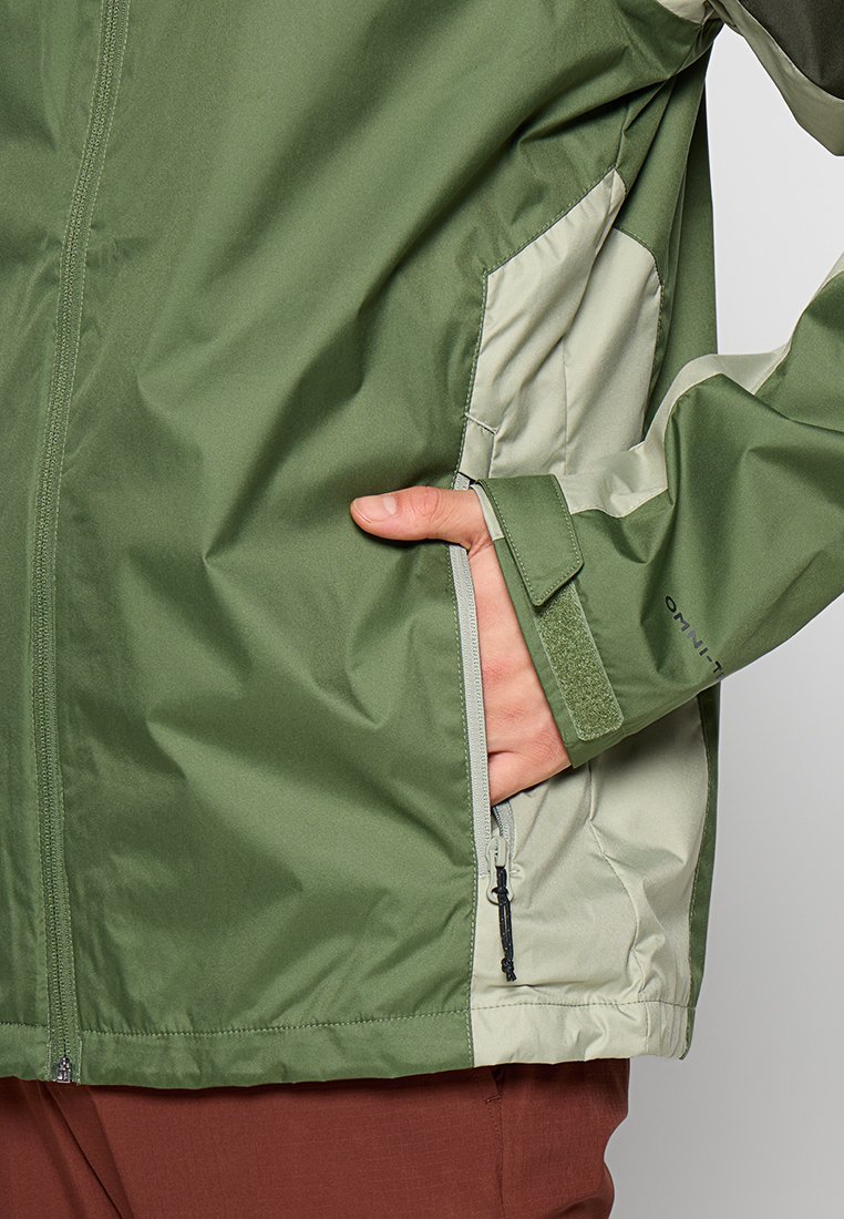 Veste verte et gris clair avec une texture lisse, fermeture éclair et fermeture Velcro. Présente une main dans la poche, mettant en avant le design et le fit.