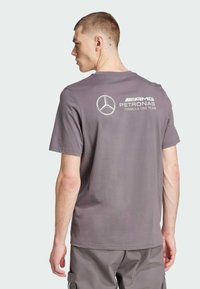 Camiseta de algodón gris con cuello redondo y mangas cortas. El diseño en la parte posterior incluye un logotipo con el emblema de Mercedes-Benz y texto.
