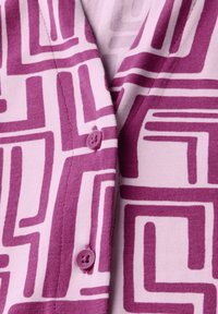 Violetti kuviollinen neuletakki, jossa on geometrinen muotoilu vaaleanpunaisella taustalla. Varustettu napeilla ja pehmeällä, neulotulla tekstuurilla.