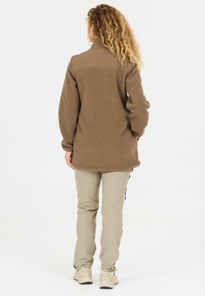 Persoon met krullend haar, gekleed in een bruin fleecejack en beige broek, met de rug naar de camera op een effen witte achtergrond.