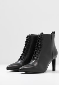 Botas de tobillo de cuero negro con punta afilada, que presentan un diseño de cordones y un tacón alto y delgado. Textura suave con un estilo minimalista.