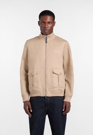 Mand iført en beige lynlåstrøje med knapbesatte lommer og mørkeblå jeans, stående foran en ensfarvet hvid baggrund.