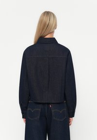 Lyöty tumman denimin takki, jossa on klassinen kaulus, napitettu hihansuu ja takana kaulus yksityiskohta. Yhdistetty korkeavyötäröisten tumman denimin housujen kanssa.