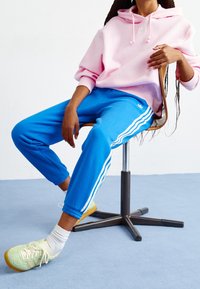 Felpa con cappuccio rosa con cordoncini, pantaloni della tuta blu con strisce laterali bianche e scarpe verde chiaro su una figura seduta su uno sfondo neutro.