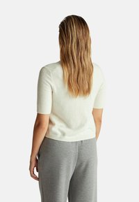 Frau mit langen, gewellten blonden Haaren, die einen kurzärmligen weißen Pullover und graue Hosen trägt, mit dem Rücken zu einer schlichten hellen Hintergrund. 