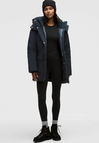Parka imbottita blu navy con cappuccio removibile, bottoni a pressione frontali e due tasche laterali, abbinata a leggins neri e stivali neri con lacci.