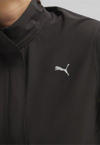 Svart träningsjacka med hög krage, slät yta och en vit Puma-logotyp på vänster bröst. Dragkedja framtill.