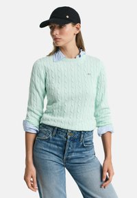 Maglione in filato di cotone verde menta con maniche lunghe, indossato sopra una camicia azzurra con colletto, abbinato a jeans in denim di colore medio; cappellino nero con dettagli dorati.