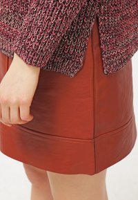 Käsi lepää kyljellä, yllään viininpuna-pinkki neulepusero ja ruosteenvärinen nahkahame, jossa näkyvät saumausvaiheet.