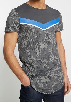 Szary T-shirt z krótkim rękawem z wzorem kamuflażu, paskiem z wzorem szewrona w jednolitym niebieskim kolorze i białymi akcentami na piersi. Zaokrąglony dół.