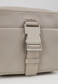 Beżowa skórzana torba o gładkiej fakturze, z metalowym klamrą w kolorze srebrnym i paskiem oraz przeszyciami na brzegach.