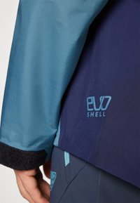 Veste bicolore avec une partie supérieure bleu clair et une partie inférieure bleu foncé. Le texte "evo Shell" est imprimé en bleu clair sur le côté.
