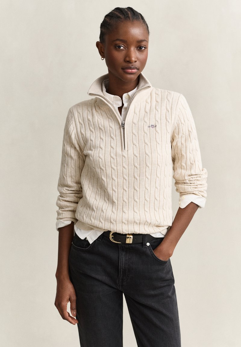 Jeune femme aux cheveux tressés porte un pull crème à demi-fermeture éclair en maille torsadée sur une chemise blanche, avec un jean noir et une ceinture noire, debout avec une main dans la poche.